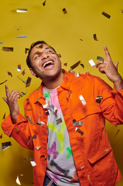 opgewonden indiaanse man in oranje jasje gebaren in de buurt confetti op gele achtergrond, party concept - Foto, afbeelding