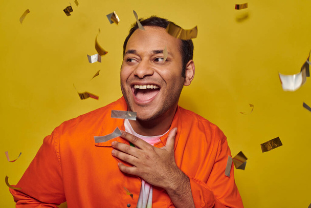 hombre indio positivo en chaqueta naranja gesto cerca de confeti sobre fondo amarillo, concepto de fiesta - Foto, Imagen