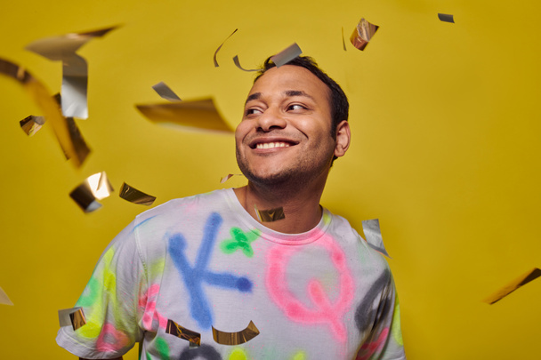 zadowolony Indianin człowiek w t-shirt uśmiecha się w pobliżu spadające konfetti na żółtym tle, koncepcja partii - Zdjęcie, obraz
