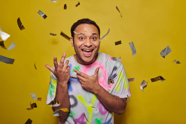 excited indian man smiling near falling confetti on yellow backdrop, party concept, merry event - Photo, Image