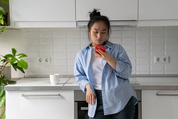 Concentrated emotionless Asian woman reading message on phone standing in kitchen. Carried away Korean girl chatting on social media is digitally addicted spending lot time on internet - Photo, Image
