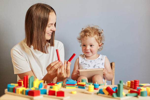 Curious child's playful learning. Wooden toys for fun. Exploring together in the nursery. Smiling mother and daughter playing learning games with wooden toys isolated over gray background - Photo, Image