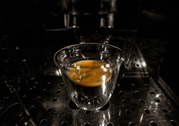 Glasschale mit frisch gebrühtem schwarzen Kaffee mit Schaum und goldener Sahne mit Blasen, die durch die Transparenz der Tasse einer vollautomatischen Espressomaschine sichtbar sind. Dunkler schwarzer und weißer Hintergrund. - Foto, Bild
