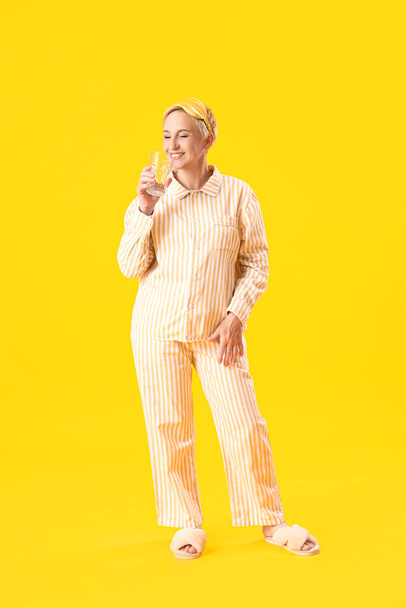Mujer madura en pijama con vaso de agua sobre fondo amarillo - Foto, imagen