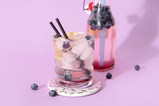 Glass and bottle of fresh blueberry lemonade on lilac background - Photo, Image