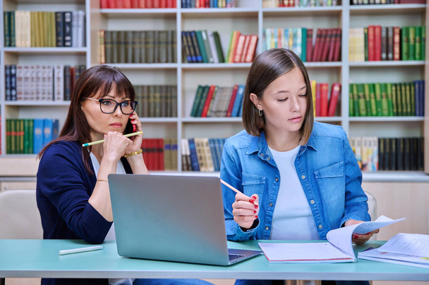 Young female university student with middle aged teacher mentor preparing for exam sitting at desk with laptop books inside library. Knowledge, education, youth, college university concept - Photo, image