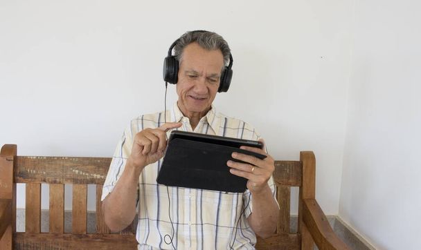 Starší muž venku sedí na lavičce a dívá se na pořad na svém iPadu Smart Tablet ve sluchátkách - Fotografie, Obrázek