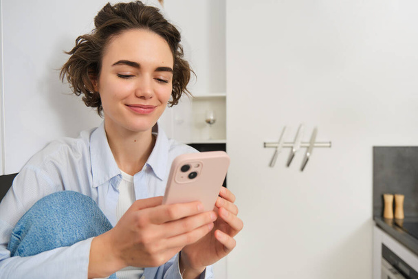 Посміхаючись брюнетка дівчина сидить зі смартфоном вдома, замовлення знімаються з мобільного додатку, використовуючи онлайн-додаток для покупок. - Фото, зображення