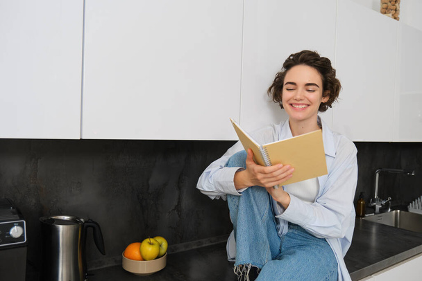 Η έννοια του τρόπου ζωής και των ανθρώπων. Νεαρή γυναίκα απολαμβάνει το Σαββατοκύριακο στο σπίτι, διαβάζοντας σημειώσεις στο ημερολόγιό της, κάθεται στον πάγκο της κουζίνας με σημειωματάριο και χαμογελά. - Φωτογραφία, εικόνα