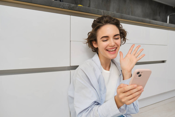 Cute brunette girl smiles at smartphone camera, waves hand, says hello, connects to video chats, talks to someone, sits on floor in kitchen. - Photo, Image