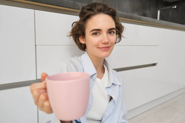 Portrait de belle fille brune, vous donne tasse avec café ou thé, offre de boire matcha avec elle, tient une tasse rose, s'assoit sur le sol de la cuisine et sourit à la caméra. - Photo, image