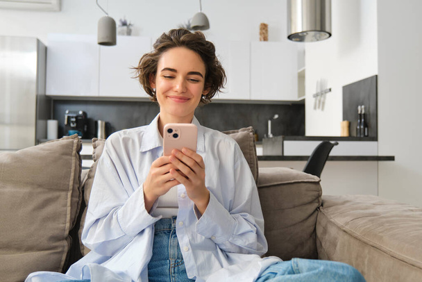 Portrait of smiling woman chats on smartphone app, sits at home on sofa and uses mobile phone application. Copy space - Photo, Image