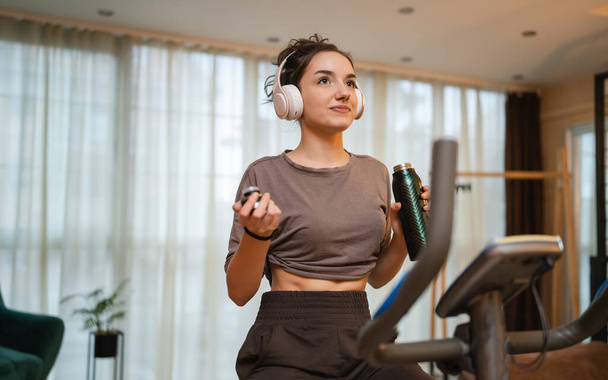 One woman young caucasian female training at home with headphones on Indoor Cycling stationary Exercise Bike real people copy space health and fitness workout hold bottle of water or supplement - Foto, imagen