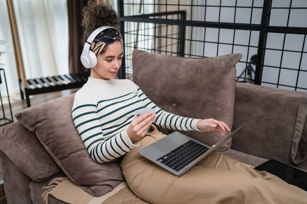 Μία γυναίκα που ψωνίζει online κατέχει πιστωτική κάρτα και φορητό υπολογιστή στο σπίτι πίσω όψη της νεαρής ενήλικης γυναίκας που κάνει online αγορά, ενώ κάθεται στον καναπέ-κρεβάτι στο σπίτι πραγματικούς ανθρώπους αντιγράψετε χώρο - Φωτογραφία, εικόνα