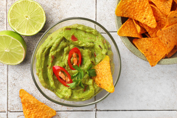 Bowl of delicious guacamole with chili pepper, nachos chips and lime on white tiled table, flat lay - 写真・画像