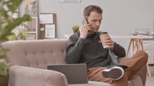 Pomalý sebevědomý muž podnikatel zamračený při telefonickém rozhovoru, sedí na gauči v kanceláři s odnášejícím šálkem kávy a notebookem - Záběry, video