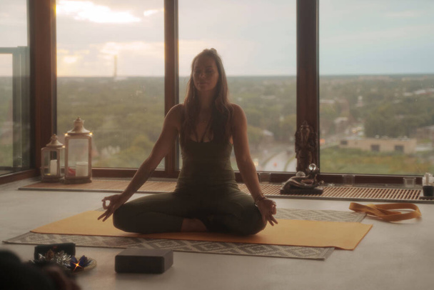 Frau in grüner Sportkleidung praktiziert Yoga und Meditation in Lotuspose vor einer panoramischen Stadtkulisse in einer Wohnung - Foto, Bild