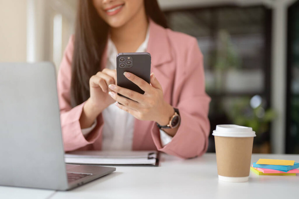 現代のオフィスで彼女の机で彼女のスマートフォンを使用しながら彼女のラップトップ画面を見ている美しいアジアのビジネスマンのクローズアップ画像. - 写真・画像