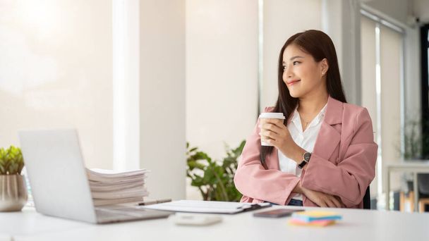 Een succesvolle en prachtige Aziatische zakenvrouw of vrouwelijke CEO in een roze business suit is het nippen van koffie, kijken uit het raam, en dagdromen over haar carrière succes aan haar bureau. - Foto, afbeelding