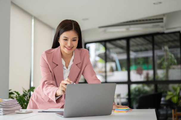Een mooie Aziatische zakenvrouw in een roze business suit buigen over een tafel en werken aan haar taken op haar laptop in het kantoor. - Foto, afbeelding