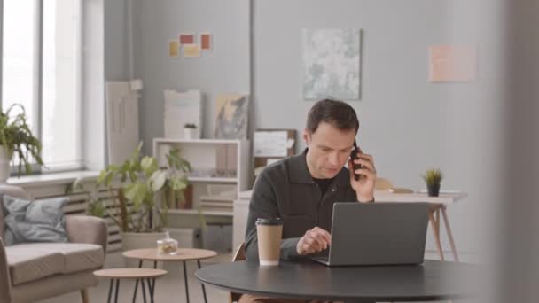 Повільність кавказького бізнесмена, який працює на ноутбуці, розмовляє на смартфоні і п'є каву з чашки кави, сидячи за зручним чорним столом в сучасному офісі - Кадри, відео