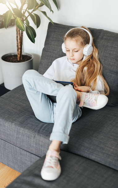 A kislány, aki a kanapén fekszik egy elektronikus táblagéppel, zenét hallgat fehér fülhallgatóban. Vezeték nélküli eszközök használata az oktatásban. - Fotó, kép