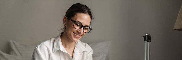 Smiling mid aged brunette white businesswoman sitting on bed in a hotel room holding mobile phone - 写真・画像