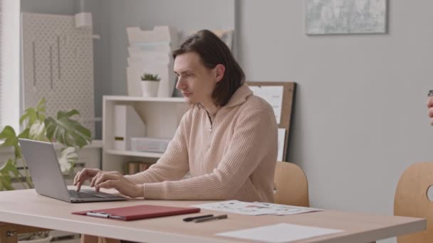 Jeune homme gay caucasien travaillant sur un ordinateur portable dans un bureau moderne tandis que son petit ami apporte du café dans des tasses à emporter pour les deux - Séquence, vidéo