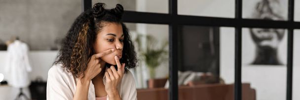 Young african woman squeezing zit while watching beauty tutorial on a laptop at home - Foto, immagini