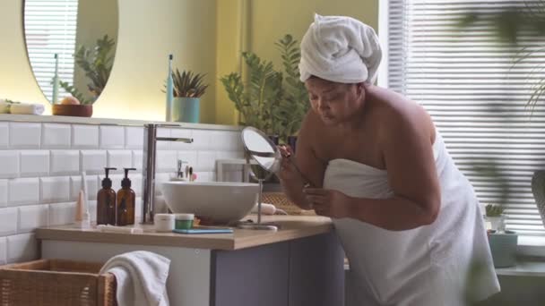 Curvy Afroamerykanka stosując tusz do rzęs podczas jej codziennego makijażu po kąpieli - Materiał filmowy, wideo