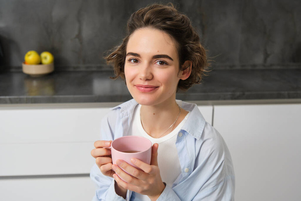 Portrait de jeune femme souriante, belle, buvant du café dans la cuisine, magie du matin avec thé cuppa, regardant tendrement. - Photo, image