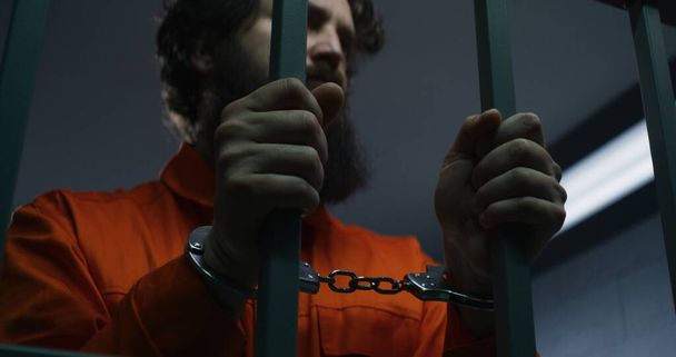 Prisoner in orange uniform and handcuffs holds metal bars, stands in prison cell. Criminal serves imprisonment term for crime in jail. Tired inmate in detention center. Justice system. Rack focus. - Photo, Image
