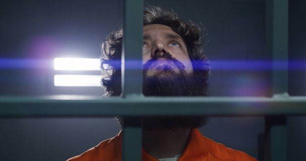 Muž v oranžové uniformě drží kovové mříže spoutanýma rukama, stojí ve vězeňské cele. Vězeň si odpykává trest odnětí svobody za zločin ve vězení. Unavený vězeň v nápravném zařízení. Zaměření regálu. - Fotografie, Obrázek