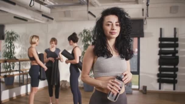 Happy Caucasian woman with curly hair holding bottle full of water and cheerfully smiling on camera. Getting ready for practicing yoga or fitness with her friends. Spending useful time. - Footage, Video