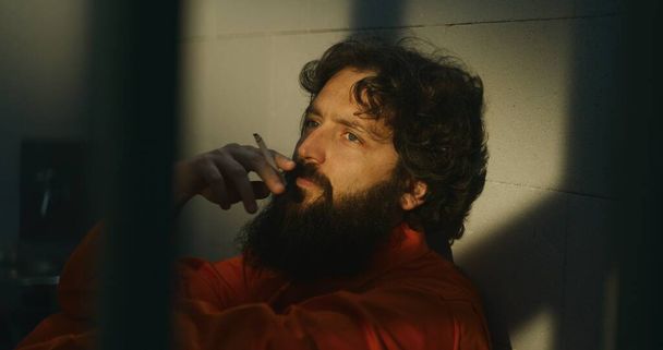 Nachdenklicher männlicher Häftling in orangefarbener Uniform sitzt auf dem Bett, zündet sich Zigarette mit Feuerzeug an und raucht in der Gefängniszelle. Strafgefangener verbüßt Haftstrafe für Verbrechen im Gefängnis. Haftanstalt. - Foto, Bild