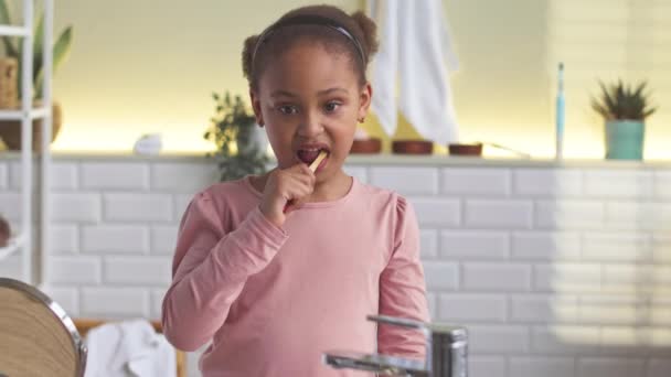 Linda niña afroamericana de 6 años mirando a la cámara mientras se cepilla los dientes en el baño por la mañana - Imágenes, Vídeo