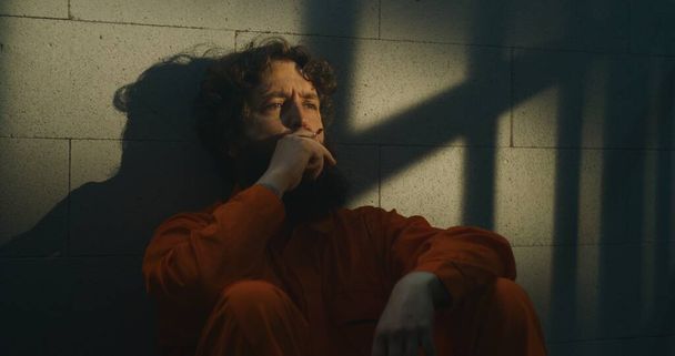 Deprimovaný vězeň v oranžové uniformě sedí na posteli a kouří cigaretu ve vězeňské cele. Trestný čin si odpykává trest odnětí svobody za zločin ve vězení. Slunce svítí skrz zamřížované okno. Ve vazebním středisku. Portrét. - Fotografie, Obrázek