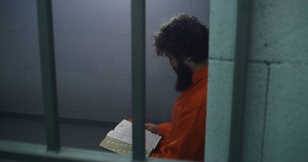 Mannelijke gevangene in oranje uniform zit op het bed, leest de bijbel, kijkt naar een raam in de cel. Crimineel zit gevangenisstraf uit voor misdaad in de gevangenis. Detentiecentrum of correctionele inrichting. - Foto, afbeelding