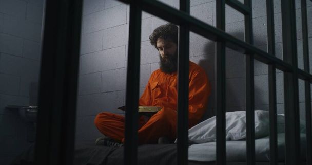 Więzień w pomarańczowym mundurze siedzi na łóżku, czyta Biblię, modli się, patrzy na zamknięte okno w celi. Więzień odsiaduje karę więzienia za przestępstwo w więzieniu. Zakład karny. Wiara w Boga. - Zdjęcie, obraz