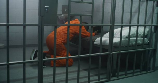 Religieuze Afro-Amerikaanse gevangene in oranje uniform knielt bij het bed, bidt tot God in gevangeniscel met bijbel. Mannelijke crimineel zit gevangenisstraf uit in de gevangenis of detentiecentrum. Geloof in God. - Foto, afbeelding