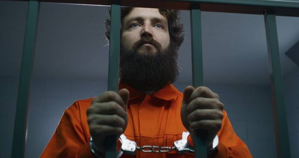 Oranssipukuinen miesvanki pitelee metallibaareja, seisoo sellissä käsiraudoissa ja katsoo kameraan. Rikollinen istuu vankeusrangaistusta vankilassa. Säilöönottokeskus tai vankila. Muotokuva. - Valokuva, kuva