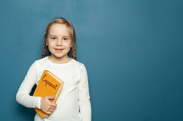 Mavi karatahtanın arka planında İspanyolca dil kitabı olan çocuk öğrenci kız - Fotoğraf, Görsel