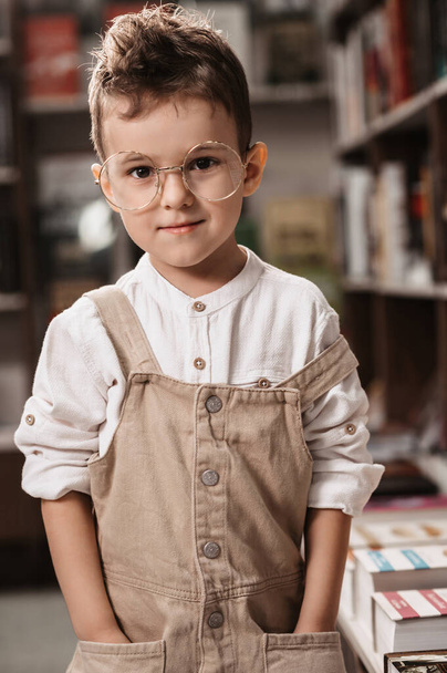 Un lindo niño caucásico, un niño con gafas redondas, en una librería entre los estantes con libros - Foto, Imagen