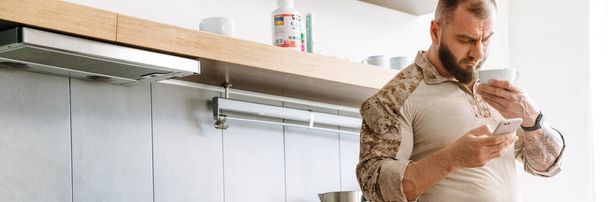 White military man using mobile phone while drinking tea in kitchen at home - Photo, image