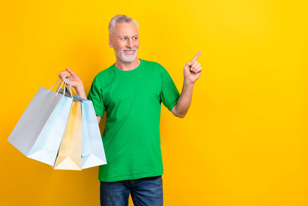 Portret van vrolijke grootvader hold winkel winkelcentrum tassen kijken direct vinger lege ruimte bieden geïsoleerd op gele kleur achtergrond. - Foto, afbeelding