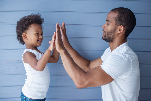 Yakışıklı genç Afro Amerikalı baba ve onun sevimli çocuğunyan görünümü elleri dokunmadan ve gülümseyerek, gri duvar önünde ayakta - Fotoğraf, Görsel