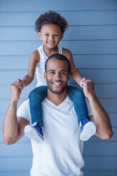 Porträt eines hübschen jungen afroamerikanischen Vaters und seines süßen Kindes, das in die Kamera blickt und lächelt. Kind sitzt auf den Schultern des Vaters - Foto, Bild