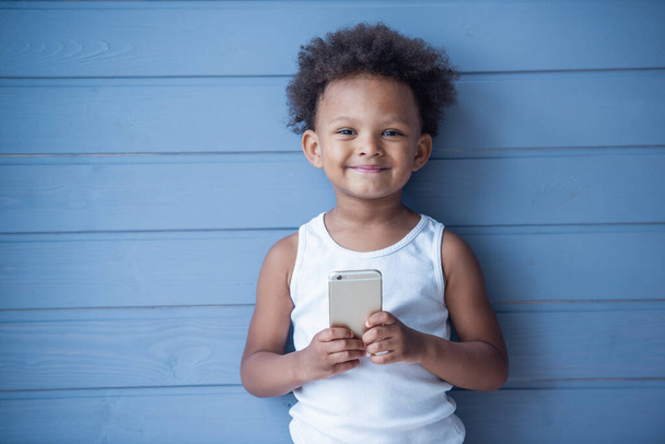 Piękna mała Afro amerykańska dziewczyna trzyma smartfon, patrząc na kamerę i uśmiechając się, stoi na szarym tle - Zdjęcie, obraz