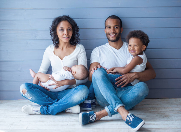 Ritratto di bei giovani genitori afro-americani con i loro figli carini stanno guardando la fotocamera e sorridendo, seduti sul pavimento contro il muro grigio. Bambino piccolo sta dormendo tra le braccia della mamma
 - Foto, immagini