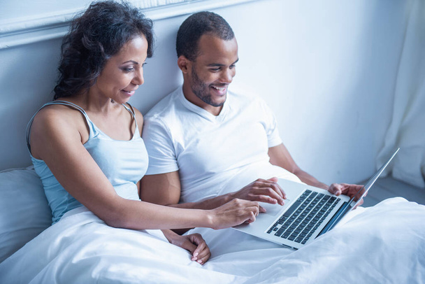 美しい若いアフロ アメリカン カップルはラップトップ コンピューターを使用し、ながら自宅のベッドで横になっている笑顔 - 写真・画像